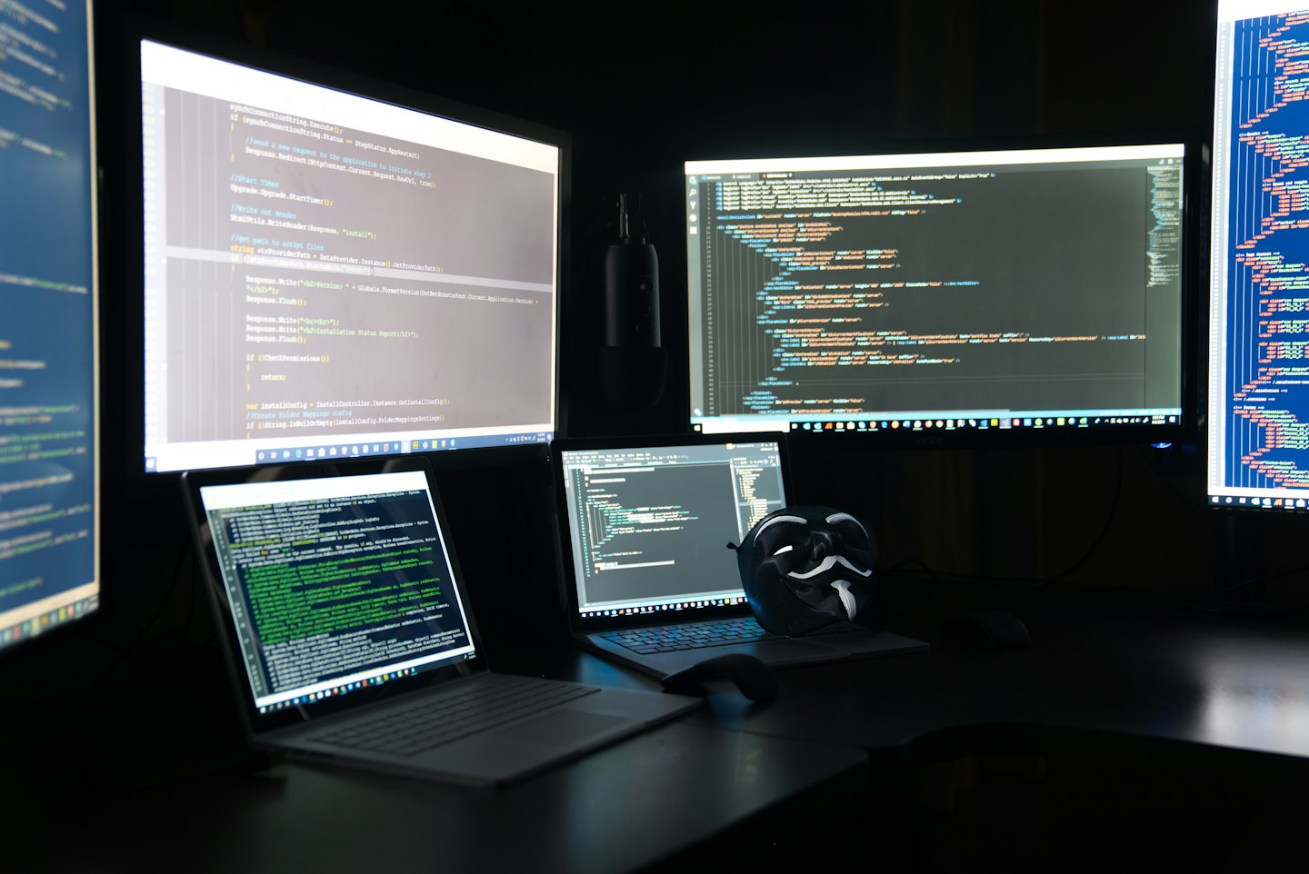 A Guy Fawkes mask resting on a keyboard among multiple monitors.